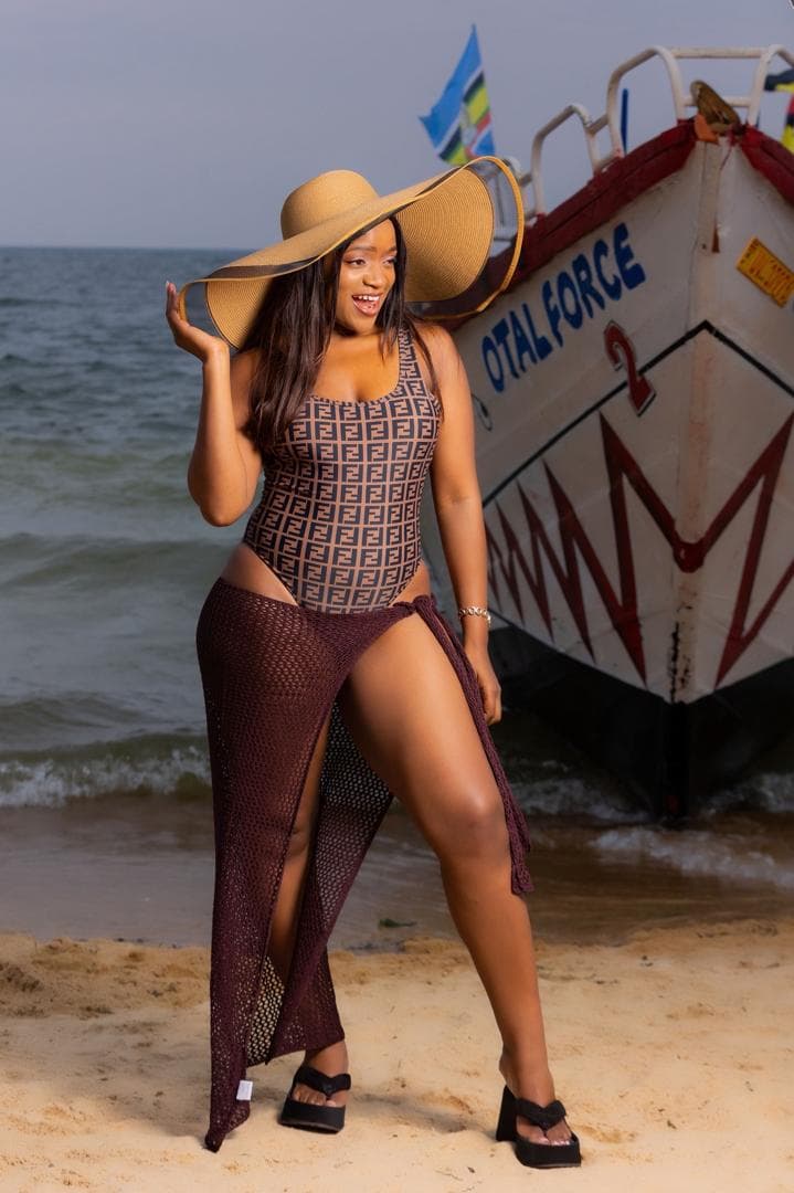 A woman in a wide-brimmed straw hat, a monogram-print one-piece and a burgundy mesh cover-up, standing on the sand beside a painted fishing boat